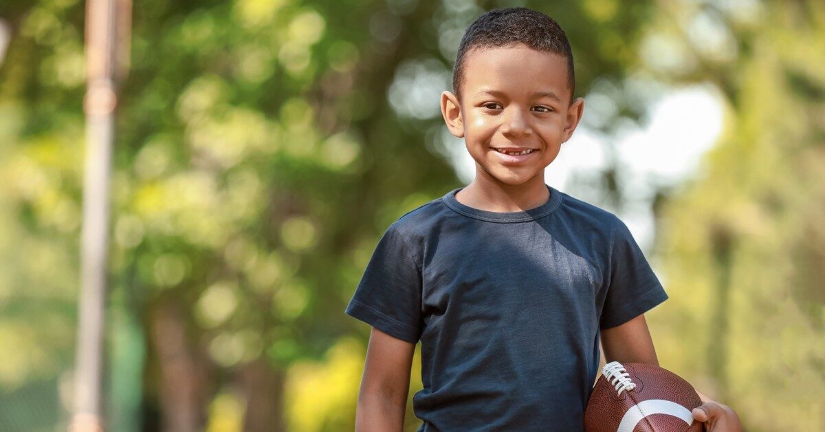 A smiling young boy wearing a black t-shirt stands in a park holding a football. Trees appear in the background.