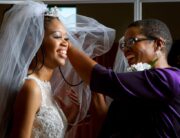 A smiling woman wearing a white bridal gown and veil. Another woman smiles as she adjusts the bride's veil.