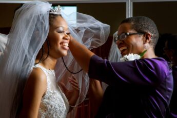 A smiling woman wearing a white bridal gown and veil. Another woman smiles as she adjusts the bride's veil.