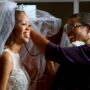 A smiling woman wearing a white bridal gown and veil. Another woman smiles as she adjusts the bride's veil.