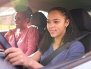 A smiling young girl is sitting behind the wheel of a car. An older woman is sitting next to her in the passenger seat.