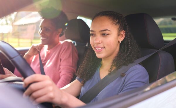 A smiling young girl is sitting behind the wheel of a car. An older woman is sitting next to her in the passenger seat.