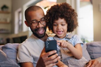 A smiling young girl sits on a man's lap on the couch, pointing and looking at the smartphone in his hand.