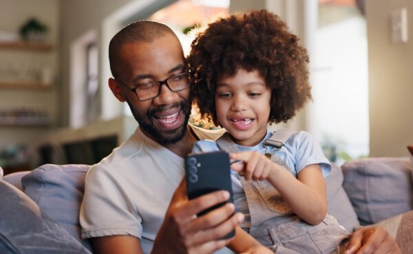 A smiling young girl sits on a man's lap on the couch, pointing and looking at the smartphone in his hand.
