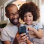 A smiling young girl sits on a man's lap on the couch, pointing and looking at the smartphone in his hand.