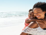 A young Black woman carrying a smiling child on her back while walking on a beach with the ocean behind her.