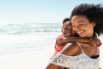 A young Black woman carrying a smiling child on her back while walking on a beach with the ocean behind her.