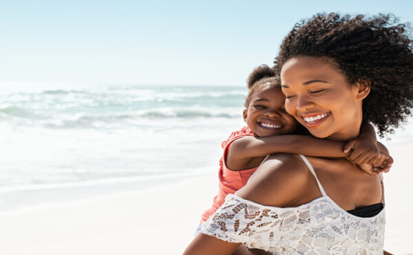 A young Black woman carrying a smiling child on her back while walking on a beach with the ocean behind her.