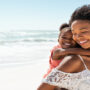 A young Black woman carrying a smiling child on her back while walking on a beach with the ocean behind her.