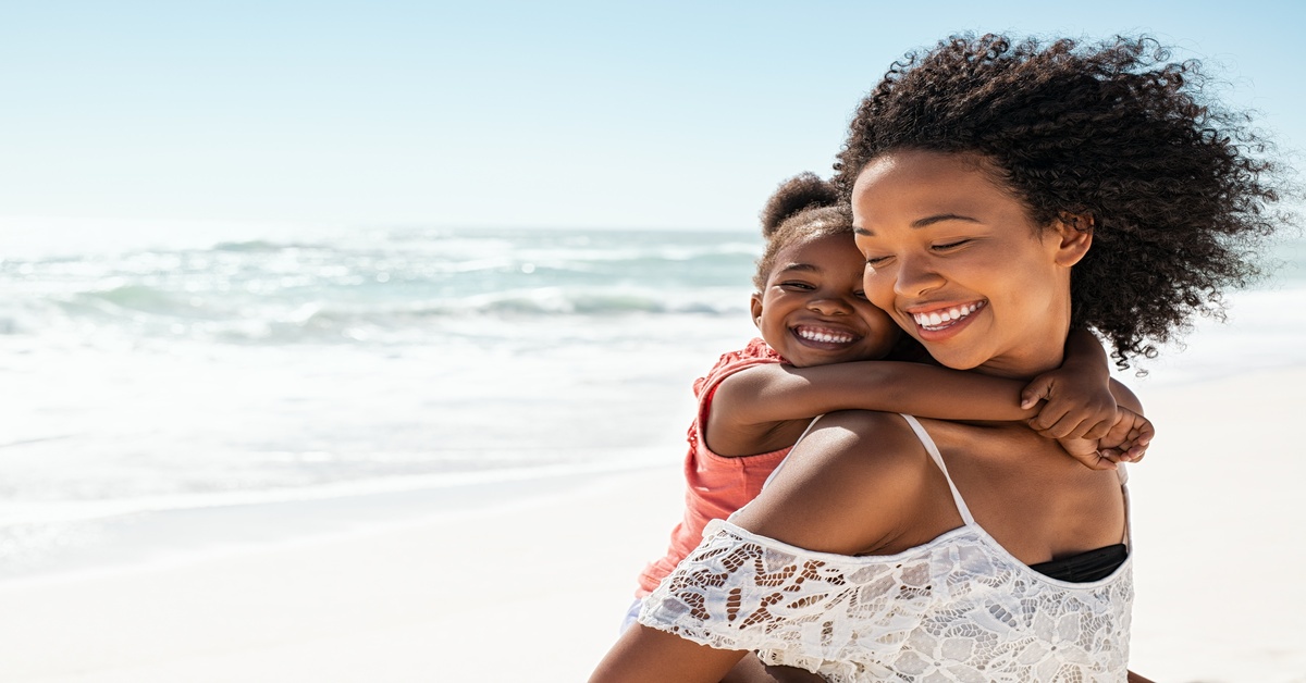 A young Black woman carrying a smiling child on her back while walking on a beach with the ocean behind her.