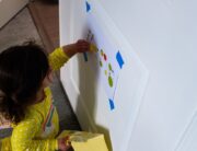 A toddler girl playing on the door with a taped sheet of paper and a sleeve of stickers. She's in a yellow outfit.