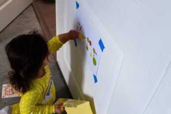A toddler girl playing on the door with a taped sheet of paper and a sleeve of stickers. She's in a yellow outfit.
