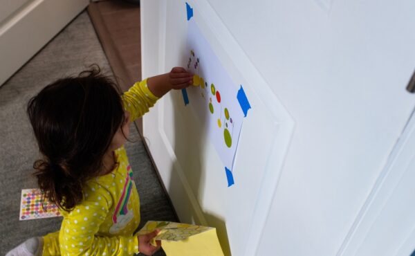 A toddler girl playing on the door with a taped sheet of paper and a sleeve of stickers. She's in a yellow outfit.