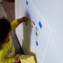 A toddler girl playing on the door with a taped sheet of paper and a sleeve of stickers. She's in a yellow outfit.