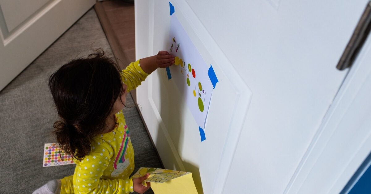 A toddler girl playing on the door with a taped sheet of paper and a sleeve of stickers. She's in a yellow outfit.