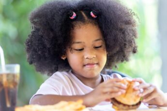 A cute Black girl sitting at a picnic table outside enjoying a hamburger, French fries, and a soda in a glass.