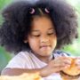 A cute Black girl sitting at a picnic table outside enjoying a hamburger, French fries, and a soda in a glass.