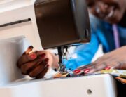 A Black woman smiles as she operates a sewing machine. She's sewing vibrant pieces of fabric together.