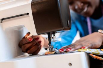 A Black woman smiles as she operates a sewing machine. She's sewing vibrant pieces of fabric together.