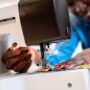 A Black woman smiles as she operates a sewing machine. She's sewing vibrant pieces of fabric together.