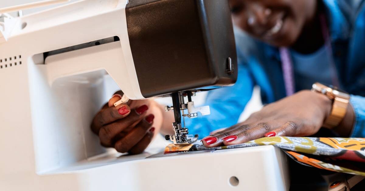 A Black woman smiles as she operates a sewing machine. She's sewing vibrant pieces of fabric together.