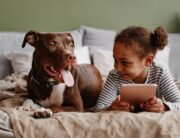 A young Black girl and a brown dog relaxing next to each other on a bed. The girl holds a tablet.