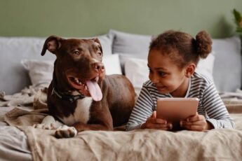 A young Black girl and a brown dog relaxing next to each other on a bed. The girl holds a tablet.