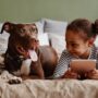 A young Black girl and a brown dog relaxing next to each other on a bed. The girl holds a tablet.