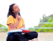 A woman wearing a yellow shirt, black pants, and sandals sits outside with a book on her lap and a pencil in her hand.