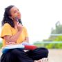 A woman wearing a yellow shirt, black pants, and sandals sits outside with a book on her lap and a pencil in her hand.