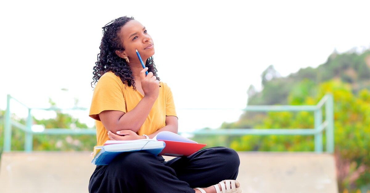 A woman wearing a yellow shirt, black pants, and sandals sits outside with a book on her lap and a pencil in her hand.