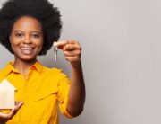 A woman in a yellow shirt smiles as she holds a key in her left hand and a wooden model house in her right hand.
