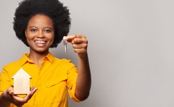 A woman in a yellow shirt smiles as she holds a key in her left hand and a wooden model house in her right hand.