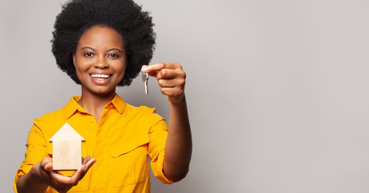 A woman in a yellow shirt smiles as she holds a key in her left hand and a wooden model house in her right hand.
