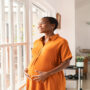 A Black pregnant woman, wearing a mustard-yellow dress, stands happily in her living room, rubbing her belly.