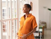 A mature Black pregnant woman, wearing an orange maxi dress, is smiling while looking out the window.