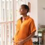 A mature Black pregnant woman, wearing an orange maxi dress, is smiling while looking out the window.