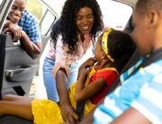 A mom smiling as she buckles in one of her kids in the backseat of a car. There is another child in the other seat.