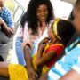 A mom smiling as she buckles in one of her kids in the backseat of a car. There is another child in the other seat.