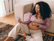 A pregnant Black woman sits on the floor of her living room, rubbing her stomach as she searches on her iPhone.