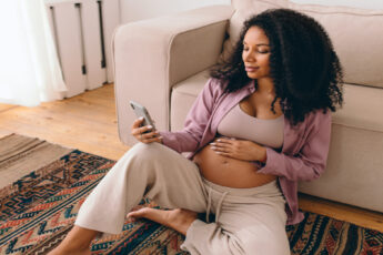 A pregnant Black woman sits on the floor of her living room, rubbing her stomach as she searches on her iPhone.