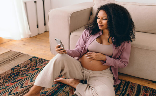 A pregnant Black woman sits on the floor of her living room, rubbing her stomach as she searches on her iPhone.
