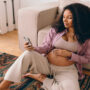A pregnant Black woman sits on the floor of her living room, rubbing her stomach as she searches on her iPhone.