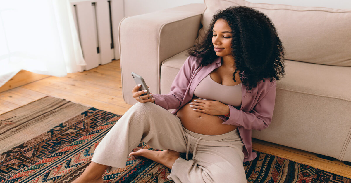 A pregnant Black woman sits on the floor of her living room, rubbing her stomach as she searches on her iPhone.