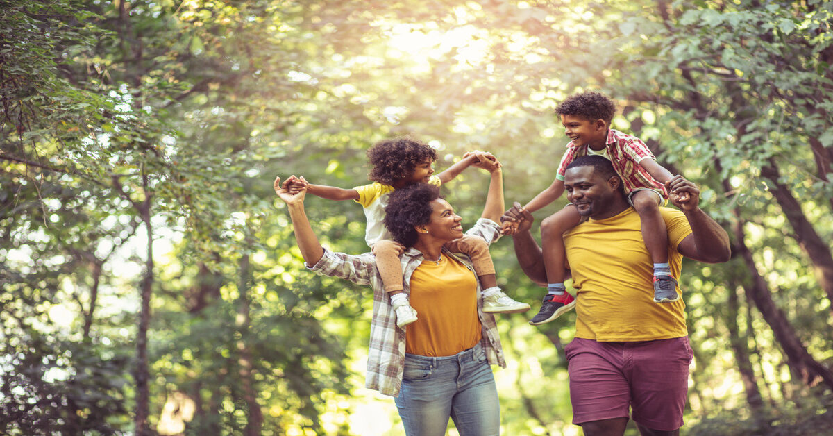 A young black family walking through the park. The parents are carrying their young children on their backs.