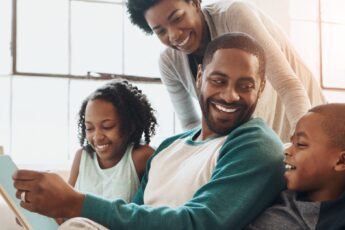 A happy Black family, a daughter, son, mom, and dad, smiles and reads together, enjoying quality time on the couch at home.