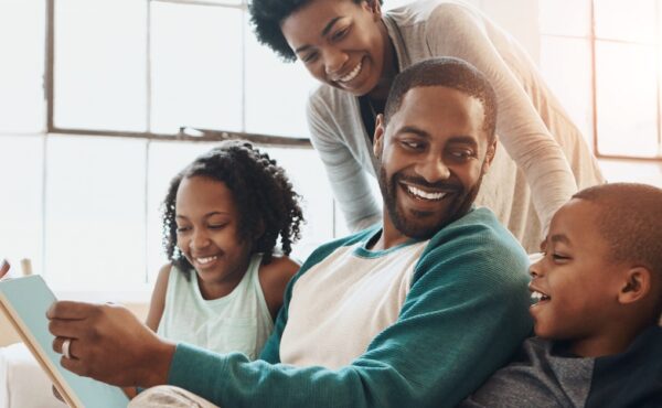 A happy Black family, a daughter, son, mom, and dad, smiles and reads together, enjoying quality time on the couch at home.