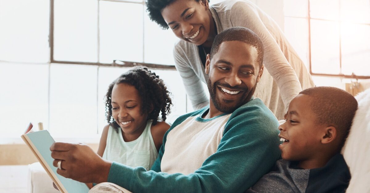 A happy Black family, a daughter, son, mom, and dad, smiles and reads together, enjoying quality time on the couch at home.