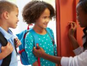 An older black woman sending off two children at the door. The kids wear backpacks and smile at the woman.