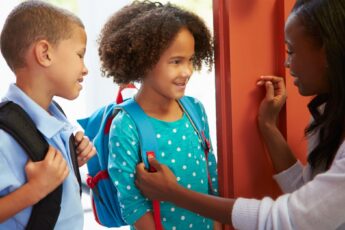 An older black woman sending off two children at the door. The kids wear backpacks and smile at the woman.